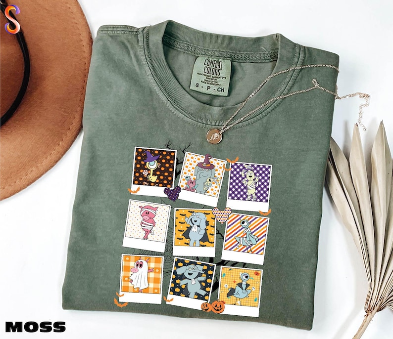 A green t-shirt with a graphic design of various cartoon characters and illustrations, including a witch, pumpkin, and other Halloween-themed elements. The t-shirt is folded and placed on a white surface, with a brown hat and some dried leaves nearby.