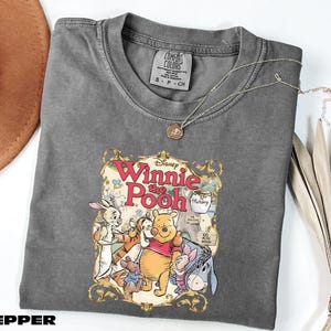 A gray t-shirt with a Disney Winnie the Pooh graphic printed on it, along with a necklace and some decorative leaves.