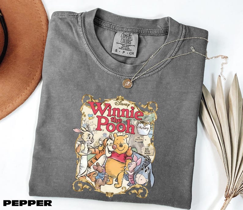 A gray t-shirt with a Disney Winnie the Pooh graphic printed on it, along with a necklace and some decorative leaves.