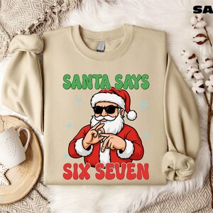 A beige sweater with a graphic design of Santa Claus and the text "Santa says six seven" printed on it, along with a snowflake and a mug.