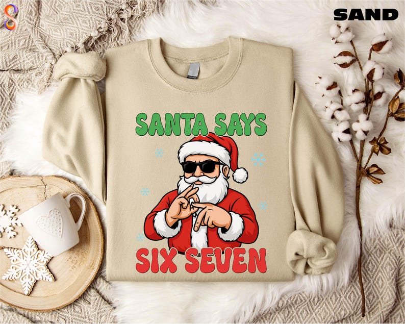 A beige sweater with a graphic design of Santa Claus and the text "Santa says six seven" printed on it, along with a snowflake and a mug.