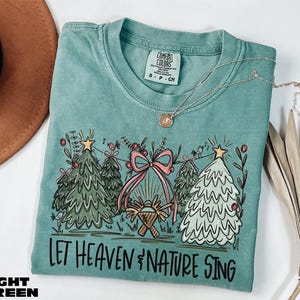 May include: Light green t-shirt with a Christmas-themed graphic. The design includes Christmas trees, a pink bow, and the text "LET HEAVEN & NATURE SING". A gold necklace is draped across the shirt.