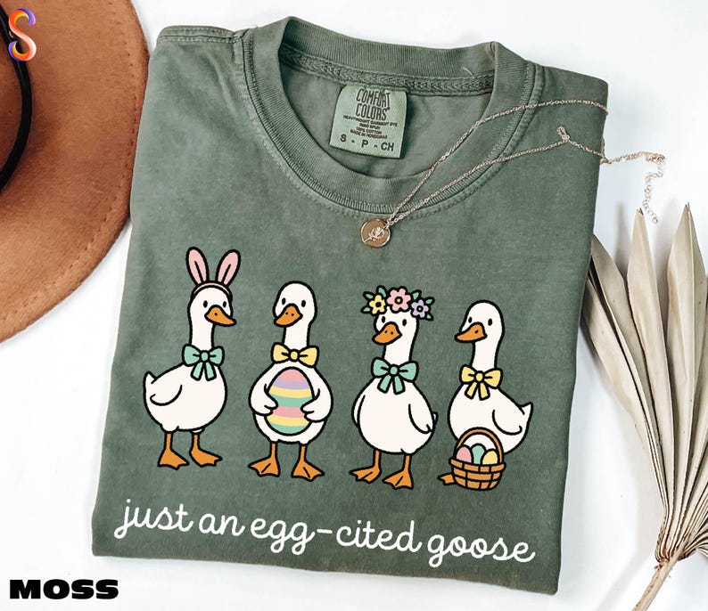 A green t-shirt with a cartoon illustration of four cartoon ducks wearing Easter-themed accessories, accompanied by the text "Just an egg-cited goose".
