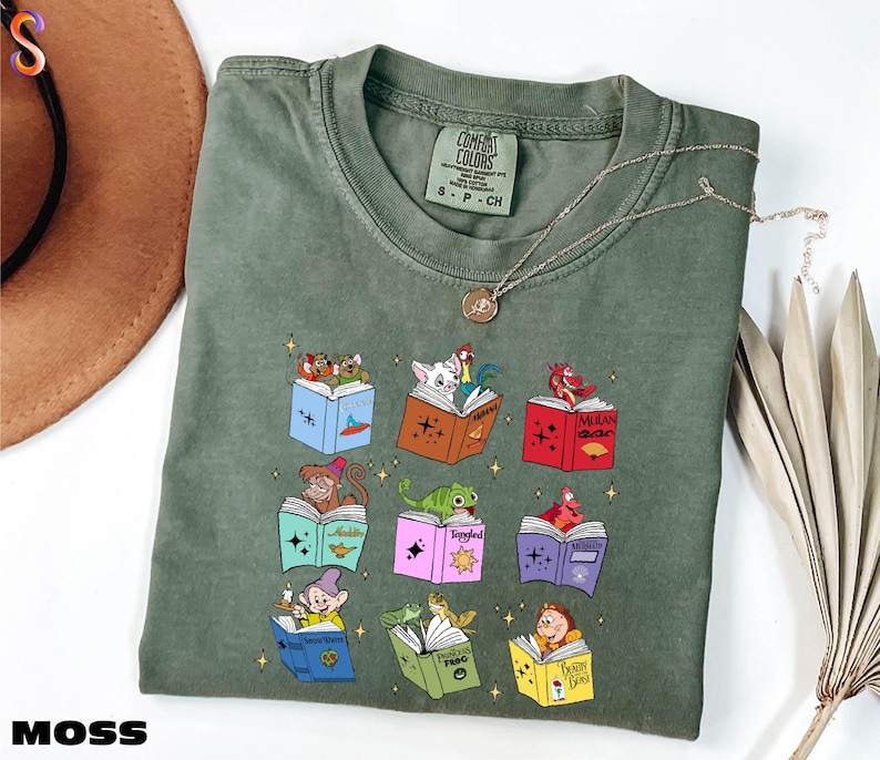 A green t-shirt with a colorful graphic of various cartoon characters and books, along with a necklace and a hat, all placed on a white surface.