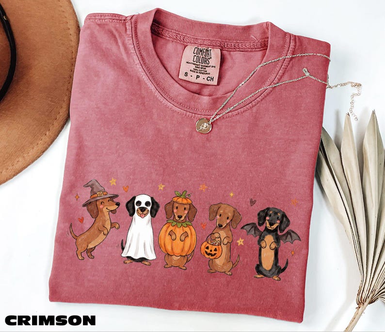 A pink t-shirt with a Halloween-themed graphic of five dogs dressed in costumes, including a witch, a pumpkin, a bat, a pumpkin, and a skeleton.
