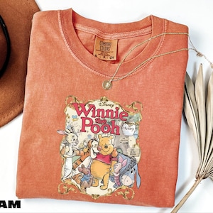 A red t-shirt with a Disney Winnie the Pooh graphic, along with a necklace and a fan, is displayed on a white surface.