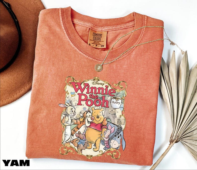 A red t-shirt with a Disney Winnie the Pooh graphic, along with a necklace and a fan, is displayed on a white surface.