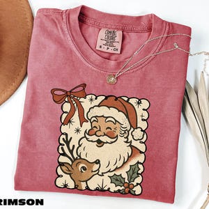 A pink t-shirt with a Santa Claus and reindeer graphic, along with a necklace and a hat, are displayed on a white surface.