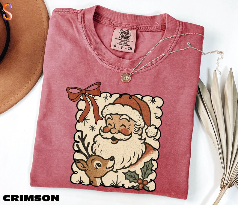 A pink t-shirt with a Santa Claus and reindeer graphic, along with a necklace and a hat, are displayed on a white surface.