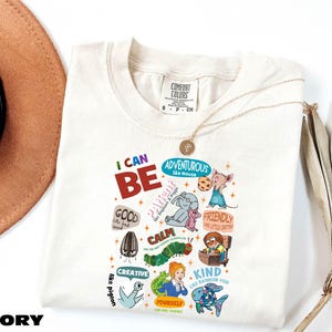 May include: Ivory-colored t-shirt with the words "I CAN BE" in red and green. The shirt features colorful illustrations of a mouse, elephant, caterpillar, and fish, along with the words "Adventurous," "Calm," and "Kind."
