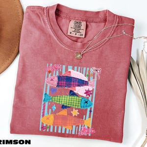 May include: A crimson short-sleeve t-shirt featuring a graphic of three stacked fish in plaid and checkered patterns. The design includes light blue vertical stripes, small stars, a heart, and a bow. The shirt has a Comfort Colors brand label.