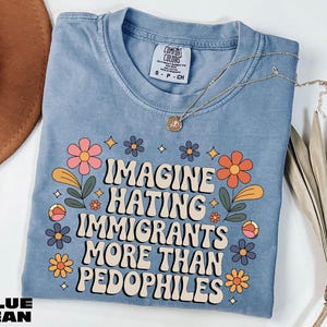 A blue t-shirt with a printed message, "IMAGINE HATING IMMIGRANTS MORE THAN PEDOPHILES", along with colorful flowers and leaves. The t-shirt is placed on a white surface, and there is a brown hat and some dried leaves nearby.