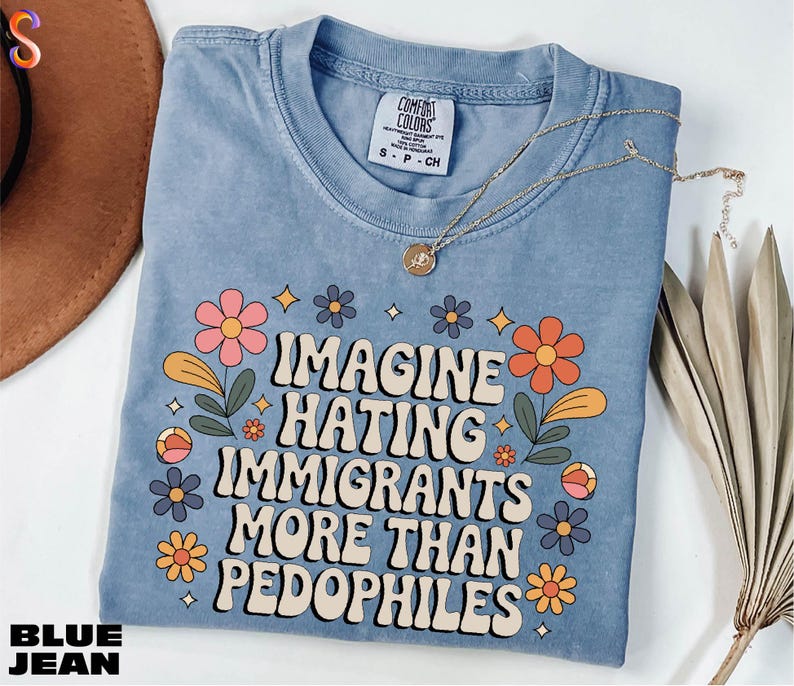 A blue t-shirt with a printed message, "IMAGINE HATING IMMIGRANTS MORE THAN PEDOPHILES", along with colorful flowers and leaves. The t-shirt is placed on a white surface, and there is a brown hat and some dried leaves nearby.