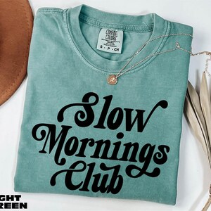 A turquoise t-shirt with the text "Slow Mornings Club" printed on it, along with a necklace and a brown hat.