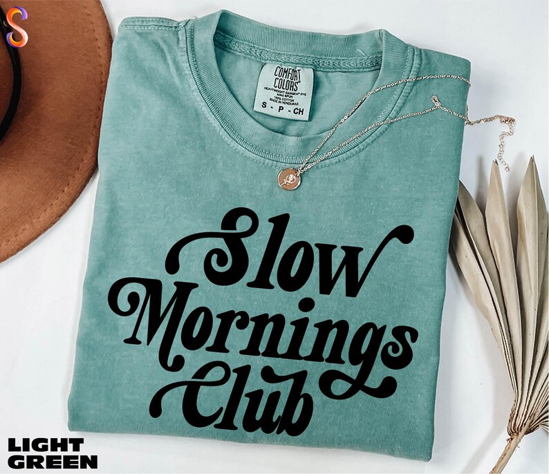 A turquoise t-shirt with the text "Slow Mornings Club" printed on it, along with a necklace and a brown hat.