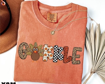 May include: A coral-colored t-shirt with the word "GOBBLE" in a playful font. The letter "G" has a leopard print, the "O" features a turkey, the "B"s have pumpkin patterns, and the "L" has a black and white checkered pattern. The "E" has a leopard print.