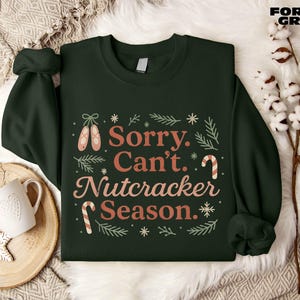 May include: Dark green sweatshirt featuring the text "Sorry. Can't. Nutcracker Season." in a festive design. The design includes ballet shoes, candy canes, and snowflakes. A white mug and snowflake decorations are visible.