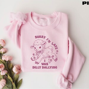 May include: Pink sweatshirt with a crew neck and long sleeves. The front features a purple graphic of a lamb with the text "SORRY I'M LATE I WAS DILLY DALLYING". The word "PINK" is in the top right corner.