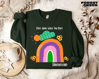 Può includere: Felpa verde foresta con un arcobaleno colorato e la scritta "Don't Judge What You Don't Understand". Il disegno include un sole e delle nuvole. La felpa è a girocollo.