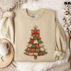 A beige sweater with a Christmas tree design printed on it, along with a mug, a pine branch, and some white cotton balls, all arranged on a white fur rug.