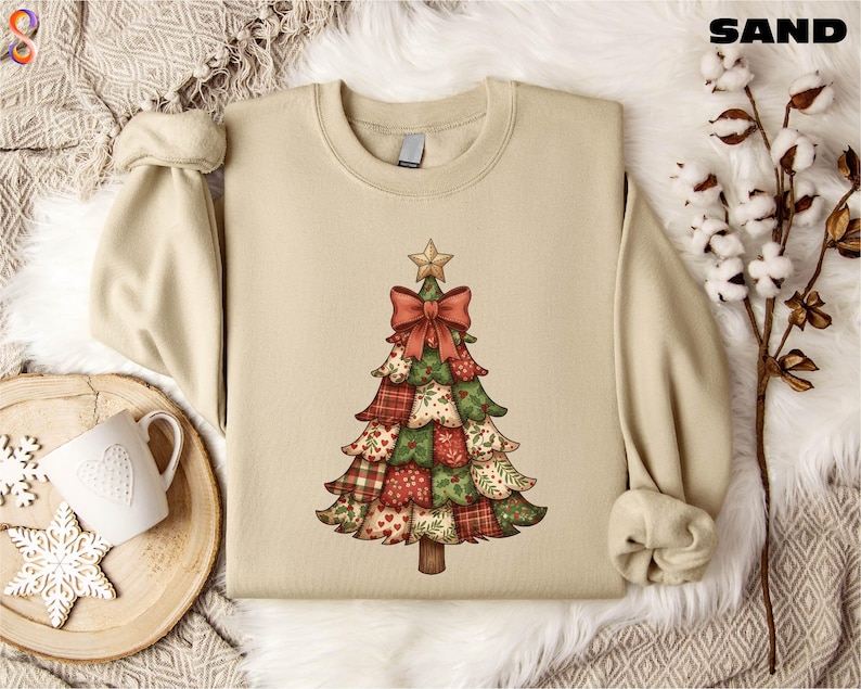 A beige sweater with a Christmas tree design printed on it, along with a mug, a pine branch, and some white cotton balls, all arranged on a white fur rug.