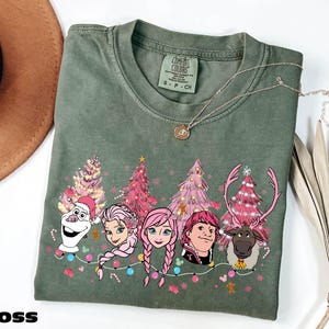 A green t-shirt with a Christmas-themed design featuring characters from the Disney movie Frozen, including Olaf, Elsa, Anna, and other popular characters.