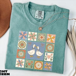 A light green t-shirt with a colorful quilt-like design featuring two ducks, flowers, and stars, along with a necklace and a hat.