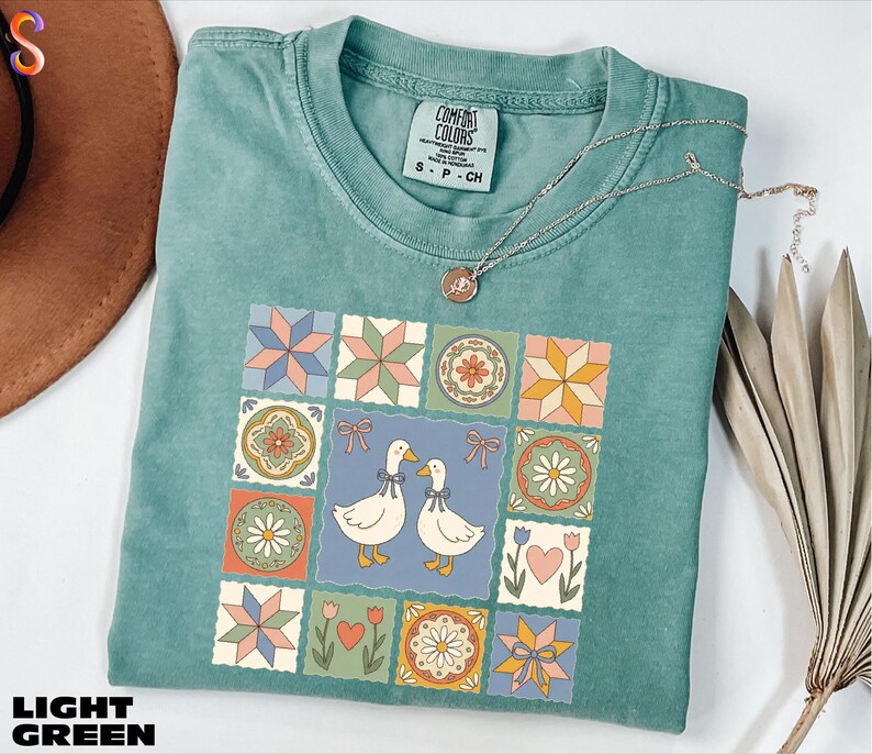 A light green t-shirt with a colorful quilt-like design featuring two ducks, flowers, and stars, along with a necklace and a hat.