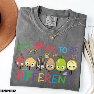 A gray t-shirt with a colorful graphic design featuring cartoon characters and the text "It's OKAY TO BE DIFFERENT" printed on it.