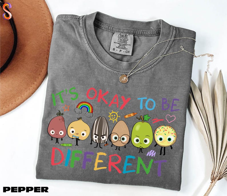 A gray t-shirt with a colorful graphic design featuring cartoon characters and the text "It's OKAY TO BE DIFFERENT" printed on it.