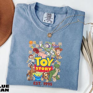A blue denim t-shirt with a graphic of characters from the Toy Story movie, including Woody, Buzz Lightyear, and other beloved characters, along with the text "Toy Story" and "Est. 1995".