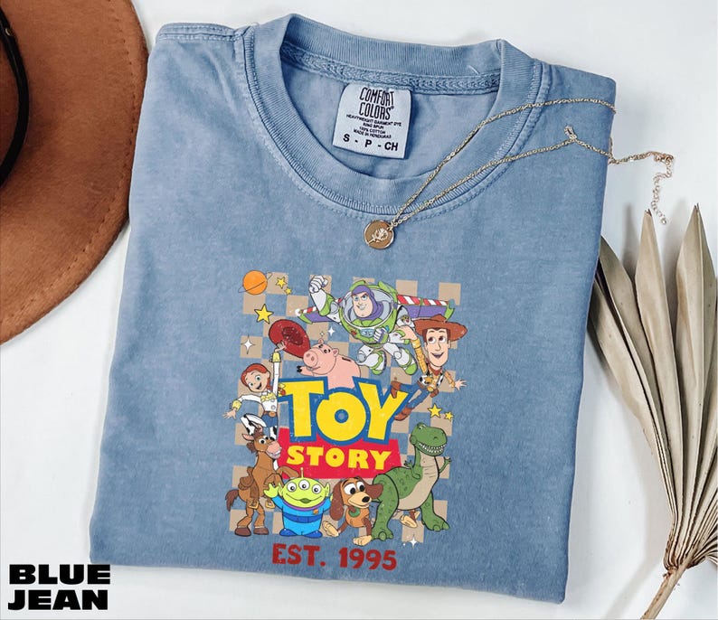 A blue denim t-shirt with a graphic of characters from the Toy Story movie, including Woody, Buzz Lightyear, and other beloved characters, along with the text "Toy Story" and "Est. 1995".