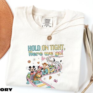 A white t-shirt with a colorful graphic of Disney characters and the text "HOLD ON TIGHT! Here we go!" printed on it, along with a necklace and a hat.