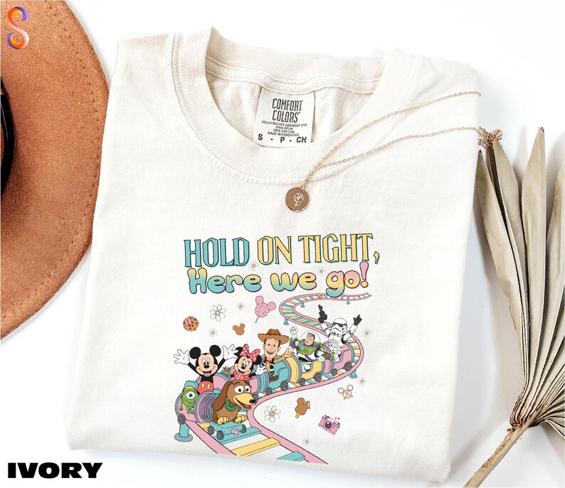 A white t-shirt with a colorful graphic of Disney characters and the text "HOLD ON TIGHT! Here we go!" printed on it, along with a necklace and a hat.