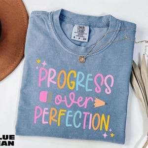 a blue t-shirt with the text "PROGRESS OVER PERFECTION" printed on it, along with a pencil graphic. The t-shirt is laid out on a white surface, and there is a brown hat and some dried leaves nearby.