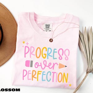 a pink t-shirt with the text "PROGRESS OVER PERFECTION" printed on it, along with a pencil and a fan. The t-shirt is laid out on a white surface, and there is a brown hat and a plant in the background.