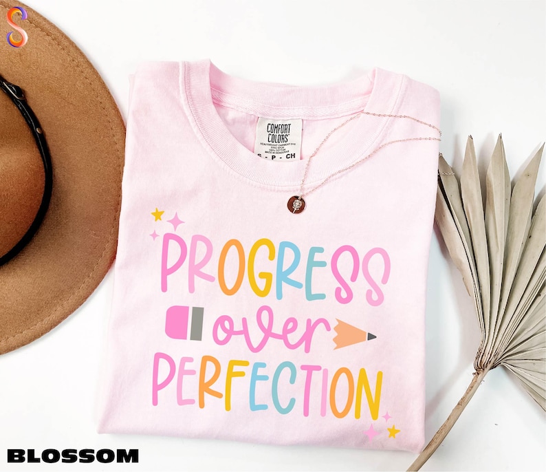 a pink t-shirt with the text "PROGRESS OVER PERFECTION" printed on it, along with a pencil and a fan. The t-shirt is laid out on a white surface, and there is a brown hat and a plant in the background.