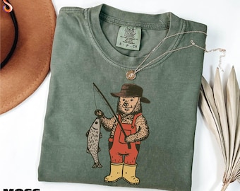 Peut inclure: T-shirt vert mousse avec un ours portant un chapeau, une salopette et des bottes, tenant une canne à pêche et un poisson. Le t-shirt a un col rond et des manches courtes. Le mot "MOSS" est imprimé en bas à gauche.