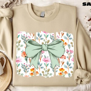 a beige sweater with a green bow on it, placed on a white fur rug. There is also a white mug and a small plant in the background.