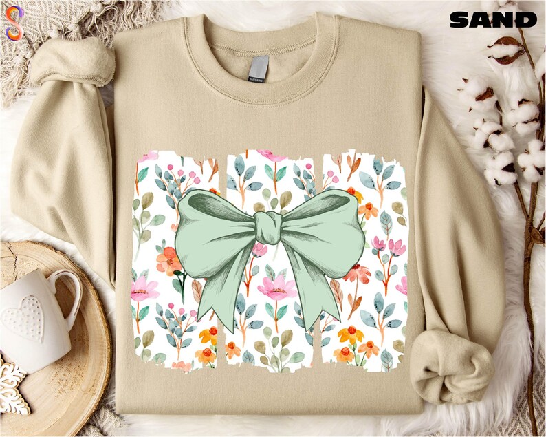 a beige sweater with a green bow on it, placed on a white fur rug. There is also a white mug and a small plant in the background.
