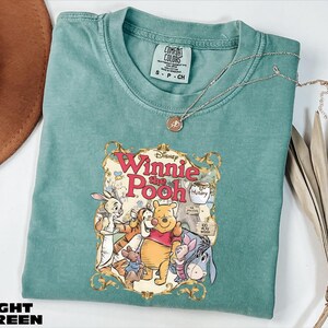 A light green t-shirt with a Winnie the Pooh graphic printed on it, along with a necklace and some decorative leaves.