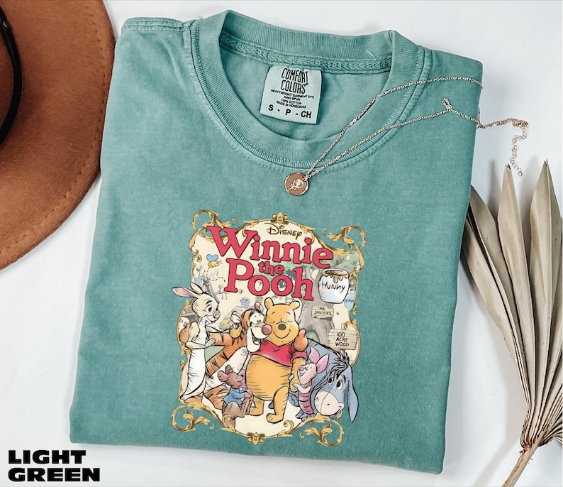 A light green t-shirt with a Winnie the Pooh graphic printed on it, along with a necklace and some decorative leaves.