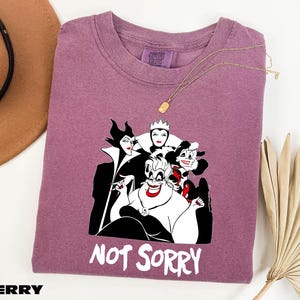 May include: Berry-colored t-shirt featuring a graphic of Disney villains, including Maleficent, the Evil Queen, and Ursula. The phrase "NOT SORRY" is printed in white. The shirt is a Comfort Colors brand.