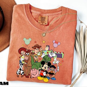 A pink t-shirt with a cartoon character design featuring characters from the Toy Story franchise, including Woody, Buzz Lightyear, and other beloved characters.