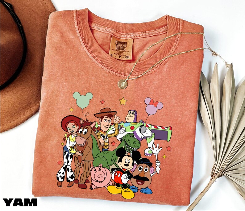 A pink t-shirt with a cartoon character design featuring characters from the Toy Story franchise, including Woody, Buzz Lightyear, and other beloved characters.