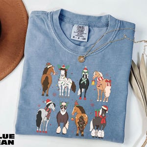 A blue t-shirt with a graphic of nine horses wearing Christmas-themed outfits, including Santa hats and stockings, is displayed on a white surface alongside a brown hat and some dried leaves.