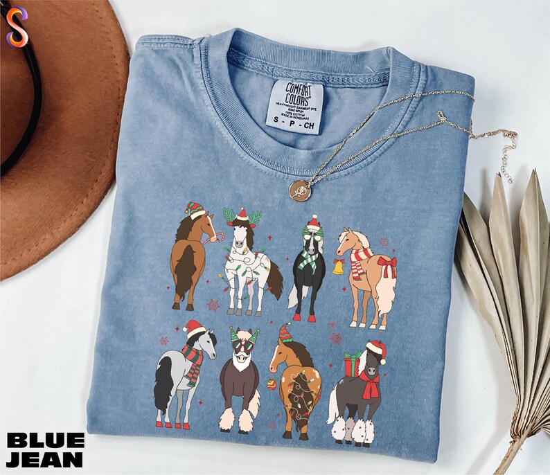 A blue t-shirt with a graphic of nine horses wearing Christmas-themed outfits, including Santa hats and stockings, is displayed on a white surface alongside a brown hat and some dried leaves.