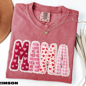 A pink t-shirt with the word "MAMA" printed on it, along with decorative heart shapes in pink and white. The t-shirt is placed on a white surface, and there is a brown hat and some leaves nearby.