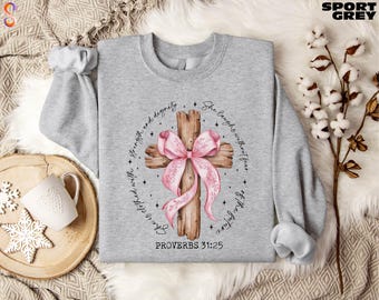 Könnte beinhalten: Ein graues Sweatshirt mit einem rosafarbenen Band, das um ein Holzkreuz gebunden ist. Der Text "Sie ist bekleidet mit Stärke und Würde. Sie lacht ohne Angst vor der Zukunft." ist um das Kreuz herum gedruckt. Der Text "Sprüche 31:25" ist unter dem Kreuz gedruckt.