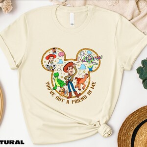 A beige t-shirt with a cartoon character design and text, along with various other items such as a hat, a woven basket, and a succulent plant.